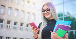 A woman looking at her phone while carrying school books.