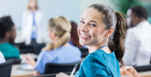 woman sitting in a classroom that's turned around in her chair and smiling at the camera. She has brown hair, in a ponytail with a blue jacket.