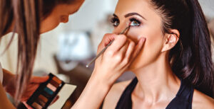 woman with her hair in a ponytail getting professional makeup done. She is looking up while the makeup artist is applying eyeliner near her eyes.