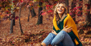 woman sitting in fall leaves, smiling and wrapped in a yellow sweater