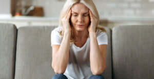 older woman sitting down with her eyes closed, she has her fingers pressed on her temples, mimicking a migraine